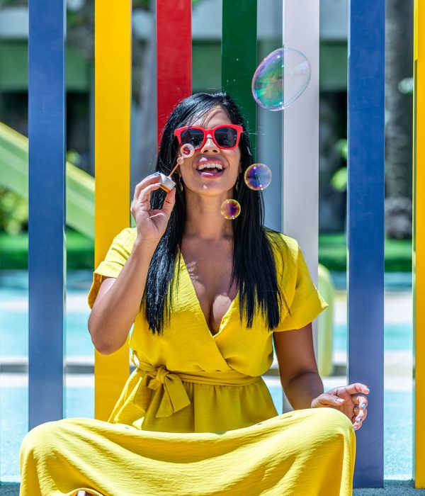 Vertical image of a Latina woman smiling and blowing bubbles soap while sitting on floor in a yellow dress against a background of a pool and colorful bars on a sunny day